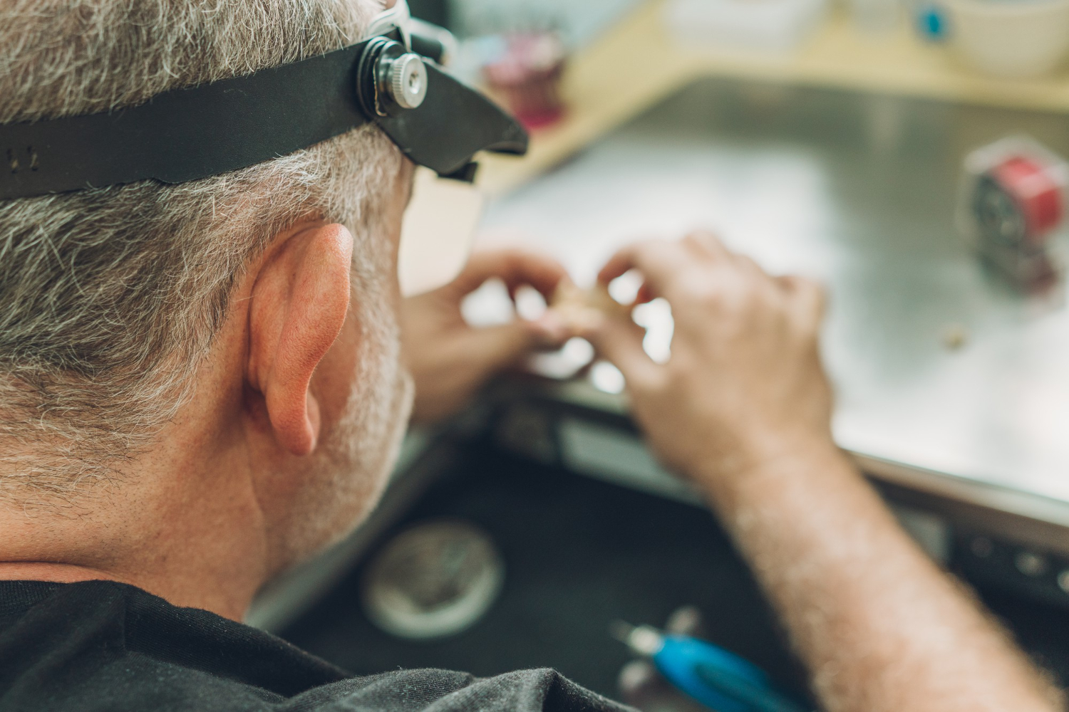 A hearing specialist in Derby wearing magnifying glasses works with small hearing aid components at a table, carefully performing detailed repair or adjustment work with specialised audiology tools nearby.