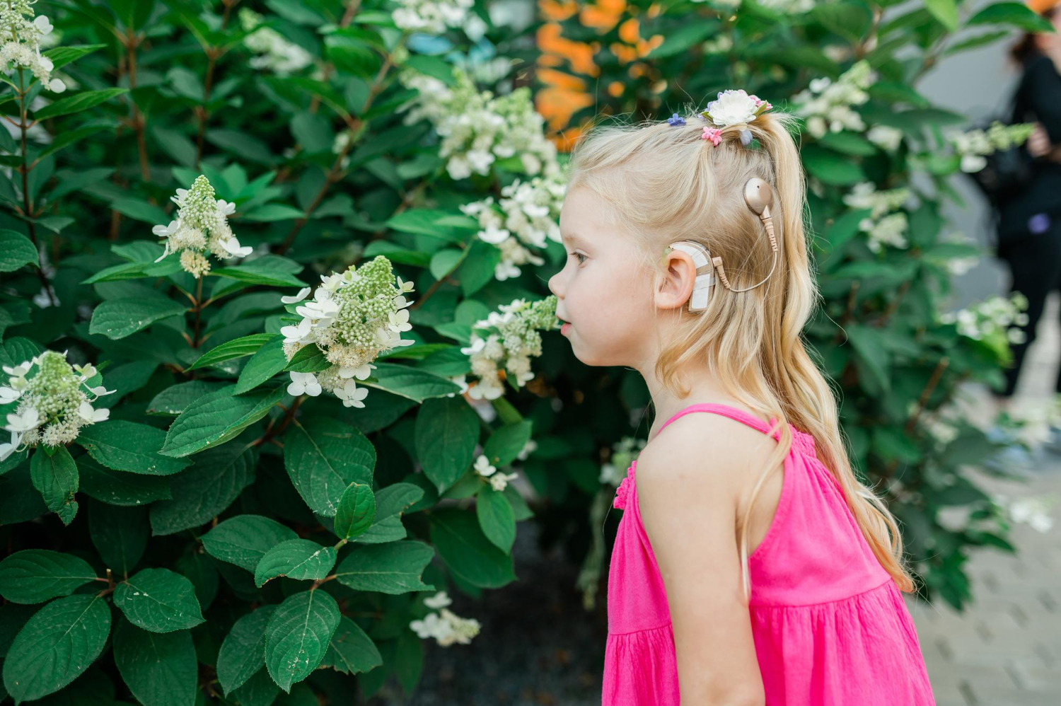 A young girl with a cochlear implant, dressed in a bright pink dress, stands beside lush green bushes adorned with white flowers, appearing to gently smell or observe the blooms&mdash;capturing a moment relevant for a Hearing Specialist in Derby.